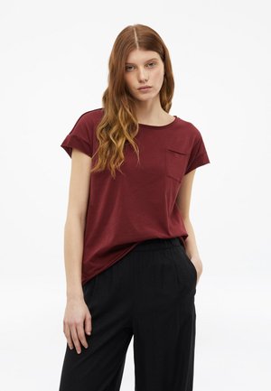 Junge Frau mit langen, welligen roten Haaren, die ein kurzärmeliges, weinrotes T-Shirt mit Brusttasche und lockere schwarze Hosen trägt, stehend vor weißem Hintergrund.