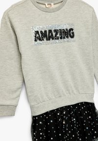 Grå sweatshirt med "AMAZING" i svarta paljetter på en silverfärgad applikation, tillsammans med en svart kjol med iriserande stjärnmönster.