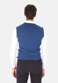 Gilet senza maniche blu in maglia con scollo rotondo, indossato sopra una camicia bianca a maniche lunghe, abbinato a pantaloni scuri, visto da dietro.