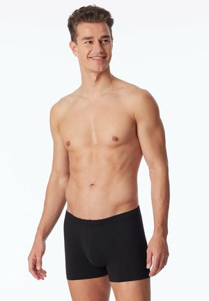 Schwarze Baumwoll-Boxershorts mit einem elastischen Taillenbund, ausgestattet mit einem körpernahen Schnitt und glatter Textur, getragen von einem männlichen Model mit neutralem Ausdruck.