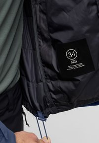 Veste rembourrée avec une couche extérieure bleue et une doublure intérieure isolante noire. Présente une étiquette détaillant le système 3-en-1 et des cordons de serrage réglables.