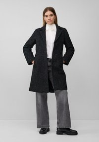 Vrouw staat met handen in de zakken en draagt een donkere, gestructureerde, knielange jas, een witte gekraagde blouse, grijze jeans en zwarte laarzen tegen een eenvoudige achtergrond.