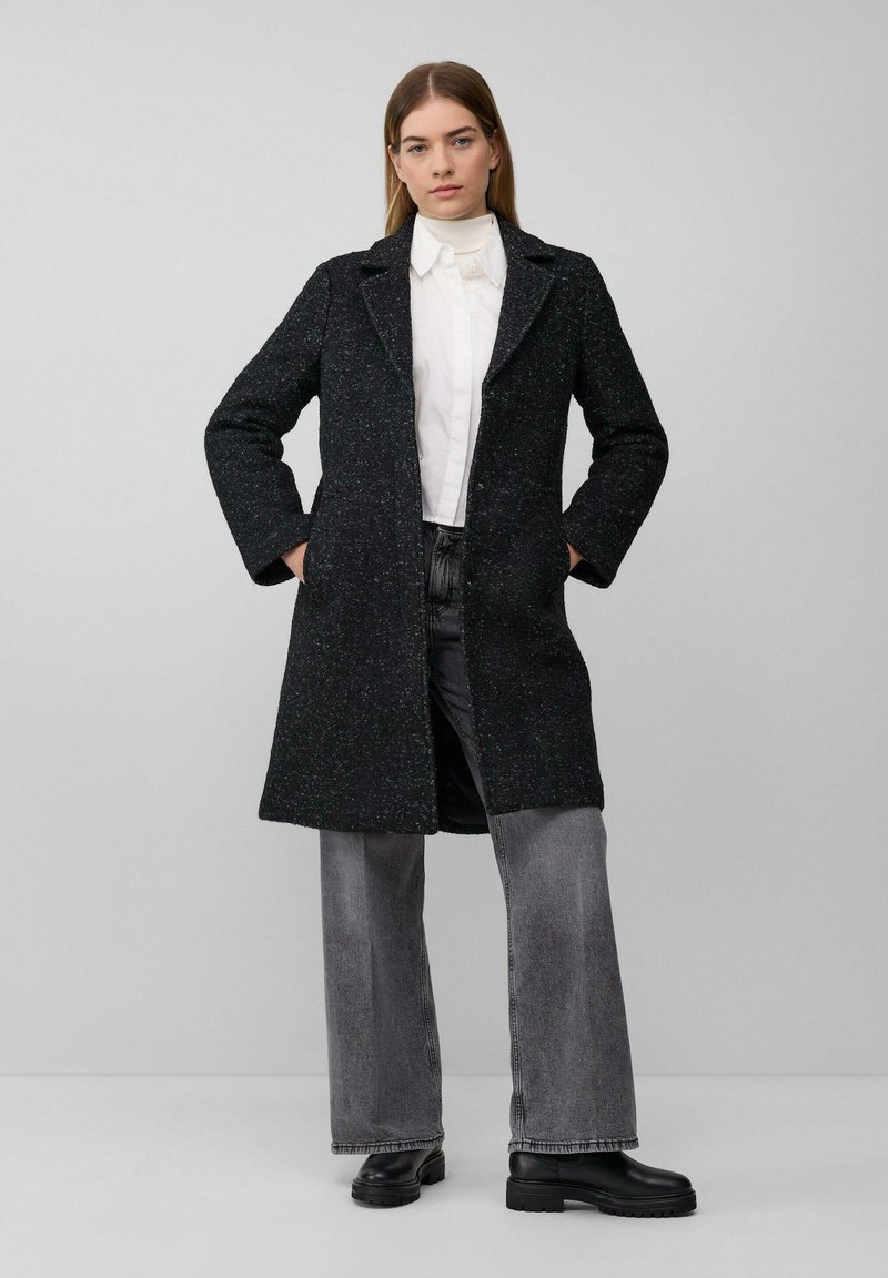 Vrouw staat met handen in de zakken en draagt een donkere, gestructureerde, knielange jas, een witte gekraagde blouse, grijze jeans en zwarte laarzen tegen een eenvoudige achtergrond.