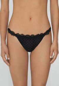 String noir avec bord en dentelle, fabriqué en tissu en maille à pois. Fini délicat et design à couverture minimale. Texture douce et légère.
