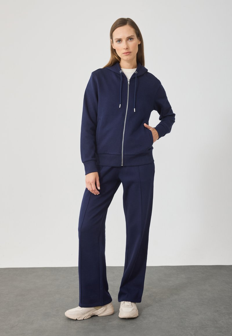 Marineblauer Reißverschluss-Hoodie und passende Hose aus einem weichen Baumwollmischgewebe. Verfügt über zwei Fronttaschen und silberne Beschläge am Reißverschluss.