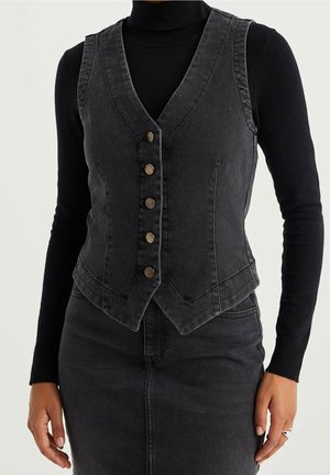 Femme portant un gilet en denim noir ajusté avec cinq boutons sur un col roulé noir à manches longues et une jupe en denim noir assortie.