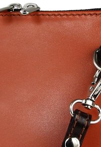 Hoek van een oranje leren handtas met een gladde textuur, voorzien van een zilveren ritssluiting en een donkerbruine riem met een metalen gesp.