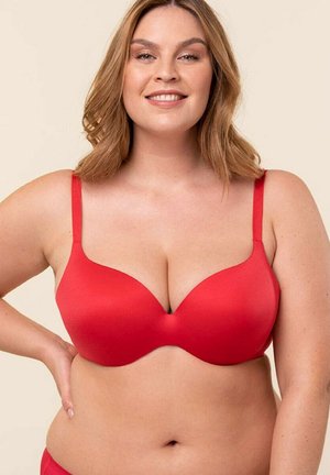 Uśmiechnięta kobieta w rozmiarze plus z blond włosami do ramion, ubrana w czerwony biustonosz na jednolitym beżowym tle.