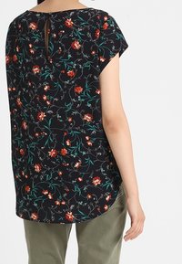 Černý květinový top s krátkými rukávy, zdobený červenými a zelenými vzory, s klíčovou zadní sponou a volným střihem, kombinovaný s olivově zelenými kalhotami.