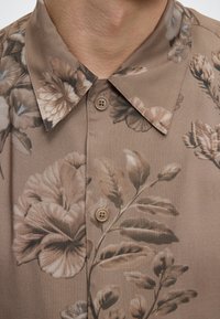 Gros plan d'une chemise marron boutonnée avec un motif floral dans des tons beiges et gris atténués et un col pointu.