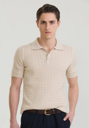 Ung mand med kort mørkt hår iført en beige kabelstrikket kortærmet polo og mørke bukser med brun bælte.