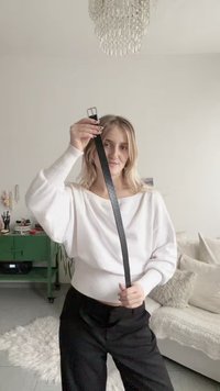 Ceinture en cuir noir avec une boucle argentée brillante, tenue horizontalement. Le modèle porte un pull blanc et un pantalon noir, debout dans une pièce aux couleurs claires.