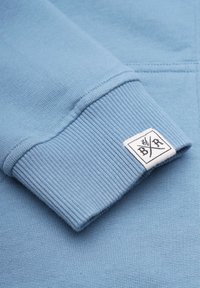 Ljust blå sweatshirt-tyg med ribbat manschett som har en liten, vit, fyrkantig etikett med svarta korsade symboler och text.