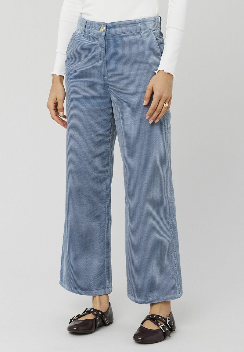 Pantalons large en velours côtelé bleu clair avec un relief prononcé, poches avant et fermeture à bouton à la taille. Portés avec des chaussures foncées.