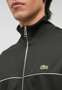 Veste noire zippée avec col montant, en tissu texturé, avec des empiècements blancs sur la poitrine et un petit logo de crocodile vert brodé.