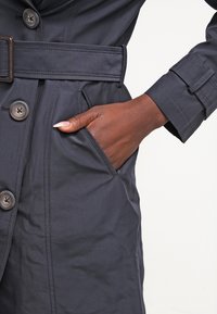 Main à la peau foncée avec de longs ongles dans la poche d'un manteau bleu marine ceinturé avec de gros boutons et des détails cousus.