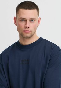 Tamsiai mėlynas džemperis su apvali apykakle, ilgomis rankovėmis ir juodu spausdintu logotipu, kuriame užrašyta "Veiksmai Sprendimai" priekyje.