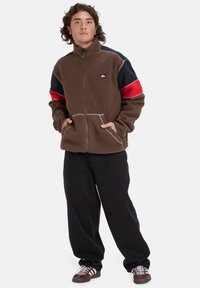 Chaqueta de forro polar marrón con detalles en negro y rojo, que cuenta con dos bolsillos frontales y un cuello alto. Lleva puesta con pantalones negros y zapatillas.