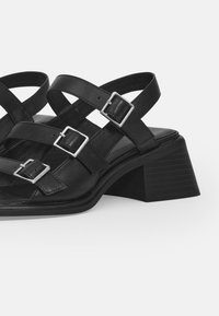 Black leather heeled sandals with multiple crisscross straps and silver buckle accents. Chunky rectangular heel with textured detailing.