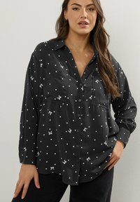 Camicia nera con bottoni, a pois bianchi e piccoli dettagli a fiocco. Presenta un colletto, due tasche sul petto e maniche lunghe. Tessuto morbido.
