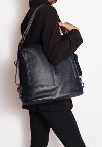 Borsa a tracolla in pelle di un blu navy scuro con superficie testurizzata, due tasche con zip e una tracolla rimovibile. Indossata sottobraccio da una persona.