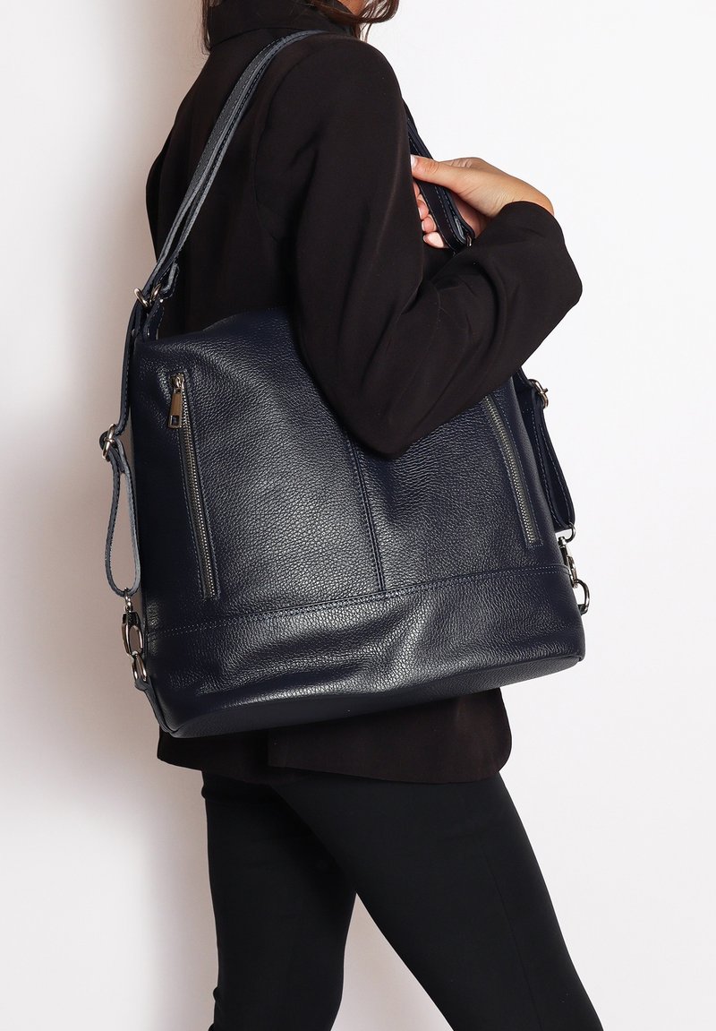 Borsa a tracolla in pelle di un blu navy scuro con superficie testurizzata, due tasche con zip e una tracolla rimovibile. Indossata sottobraccio da una persona.