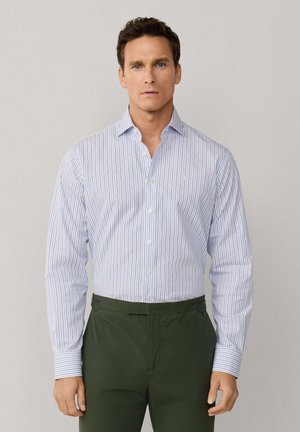 Homme portant une chemise boutonnée à rayures verticales bleu et blanc et un pantalon vert foncé, debout devant un fond clair uni.