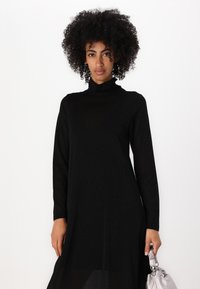 Crna haljina s turtleneck-izrezom i dugim rukavima, izrađena od glatkog materijala. Ima uski kroj i hem dužine do koljena. U paru sa srebrnom torbicom.