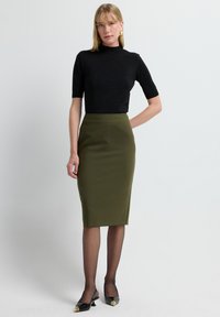 Schwarzes, eng anliegendes T-Shirt mit kurzen Ärmeln, olivgrüner Bleistiftrock, schwarze spitze Absätze mit goldenen Akzenten, transparente schwarze Strumpfhose, minimalistisches Design.