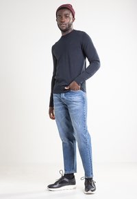 Granatowy sweter z miękkiego materiału, jasnoniebieskie jeansy i czarne trampki. Osoba ma na głowie bordową czapkę typu beanie i stoi swobodnie z jedną ręką w kieszeni.