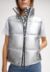 Srebrna kamizelka puchowa z wysokim kołnierzem, przednim zamkiem błyskawicznym i czarnymi detalami z logo. Gładki, błyszczący materiał z pikowanymi sekcjami.