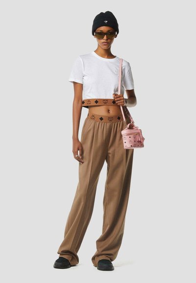 Femme portant un t-shirt blanc court, un pantalon beige avec une taille marquée par une marque, un bonnet noir, des lunettes de soleil foncées, des chaussures noires, et tenant un petit sac à main rose.