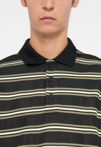 Svart og grønnstripete polo skjorte med krage, som har tre svarte knapper og et strukturert stoffmønster.