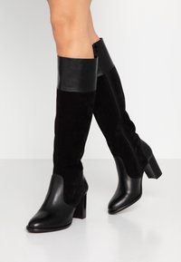 Bottes noires montantes jusqu'au genou en cuir lisse et en daim. Elles présentent un bout carré et un talon block. Fermeture éclair latérale pour un enfilage facile.