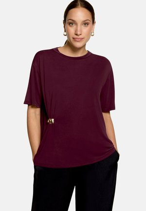 Femme à la peau claire et aux cheveux bruns portant des boucles d'oreilles créoles dorées, un haut ample à manches courtes violet foncé avec une boucle latérale, et un pantalon noir.