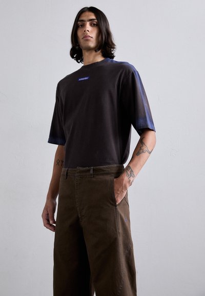 Gray oversized t-shirt with blue gradient sleeves, featuring a small blue logo near the neckline. Paired with brown, loose-fit trousers.