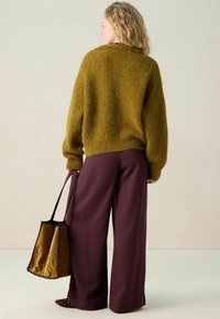 Frau in olivgrünem, kuscheligem Pullover und weiten, maronenfarbenen Hosen, die eine passende olivgrüne und maronenfarbene Tragetasche hält, mit dem Rücken zu einem schlichten Hintergrund gerichtet.