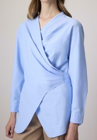 Világoskék, géz anyagú átkötős ing, V-kivágással, hosszú ujjal és aszimmetrikus alsó résszel; testhezálló fazon, puha tapintású.