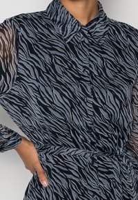 Blouse à imprimé zèbre noir et gris, dotée d'un col, d'une fermeture à boutons et de manches longues transparentes. Tissu doux avec une texture fluide.