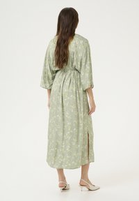 Dolga zelena cvetna obleka z zategnjenim pasom, širokimi rokavi, stranskim razporkom in lahkotnim materialom, nošena z nevtralnimi petami.