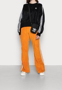 Černá manšestrová mikina s bílými pruhy, kombinovaná s jasně oranžovými manšestrovými kalhotami. Černá crossbody kabelka s bílým logem. Bílé tenisky.