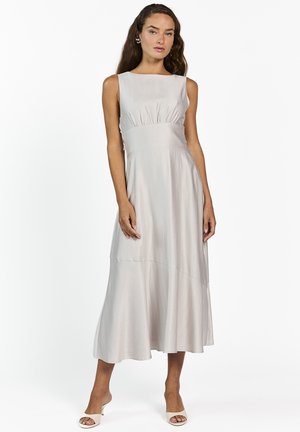 Vrouw met lang haar die een mouwloze, lichtgrijze midi-jurk en witte openhakken draagt, staand tegen een effen witte achtergrond.