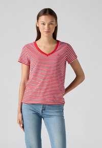 T-shirt V en jersey rouge et blanc à rayures avec des manches courtes, arborant un logo sur la poitrine gauche et un jean bleu clair. Tissu doux, design décontracté.