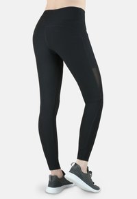 Schwarze Leggings aus dehnbarem Stoff, mit hohem Bund und Netzakzenten an den Seiten. Kombiniert mit grauen Sportschuhen.