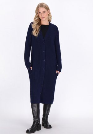 Lungo cardigan navy realizzato in tessuto a costine, con scollo a V, bottoni anteriori e tasche laterali. Indossato sopra un top nero.
