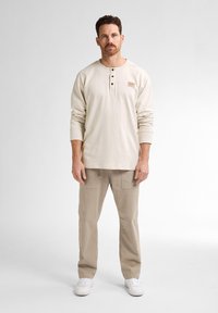 Ljusbeige långärmad henley-tröja med tre knappar och en liten etikett, kombinerat med ljus khaki cargo-byxor och vita sneakers.