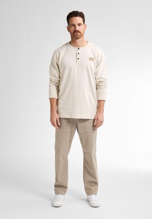 Helles beige Langarm-Henley-Shirt mit drei Knöpfen und einem kleinen Etiketten-Patch, kombiniert mit hellkhakifarbenen Cargo-Hosen und weißen Sneakers.