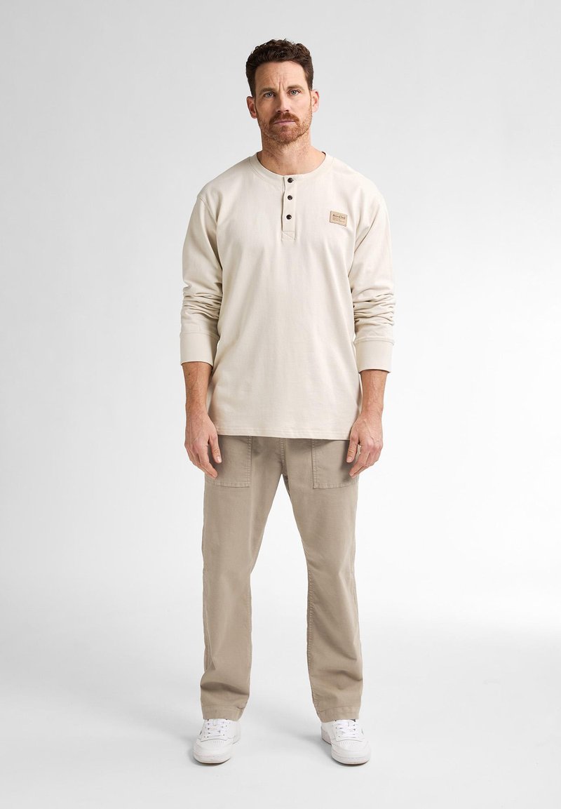 Ljusbeige långärmad henley-tröja med tre knappar och en liten etikett, kombinerat med ljus khaki cargo-byxor och vita sneakers.