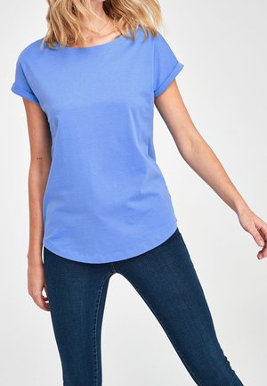 Femme portant un t-shirt bleu clair à manches courtes retroussées et un jean skinny bleu foncé sur un fond blanc.