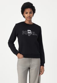 Črna sweatshirt z okroglim izrezom, ki vsebuje risani motiv in besedo "KARL" v belem. Dolgi rokavi in rebrasti robovi.